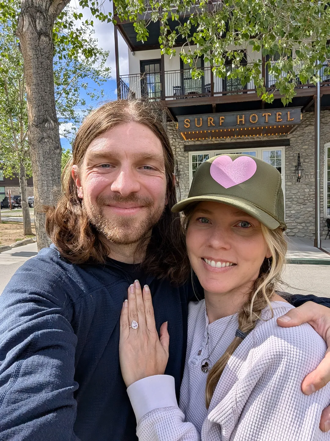 Lacy and Garrett smiling at the camera in front of the Surf Hotel, Lacy's hand resting on Garrett's chest showing her engagement ring.